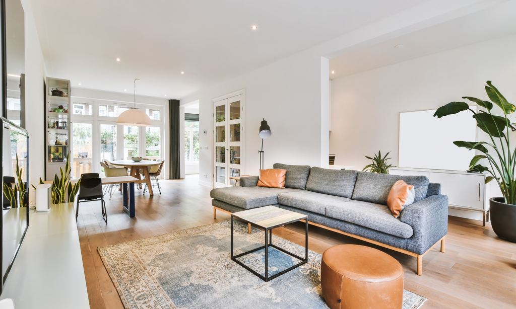 Salon moderne spacieux avec canapé gris, table basse en bois, pouf cognac, plantes vertes et grandes baies vitrées