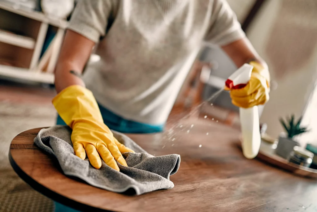 Personne réalisant le nettoyage d’une location courte durée avec produit désinfectant et chiffon sur une table en bois